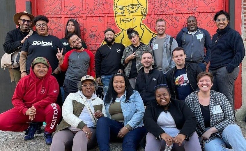 A photo at our first retreat -- a group of 16 LGBTQ+ collective volunteers, many smiling widely or displaying 'peace' signs, pose in front of a red and yellow mural of Gloria Casarez.
